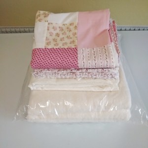 Quilt batting, muslin backing fabric, patterned binding fabric, and a finished patchwork quilt top all folded and neatly stacked