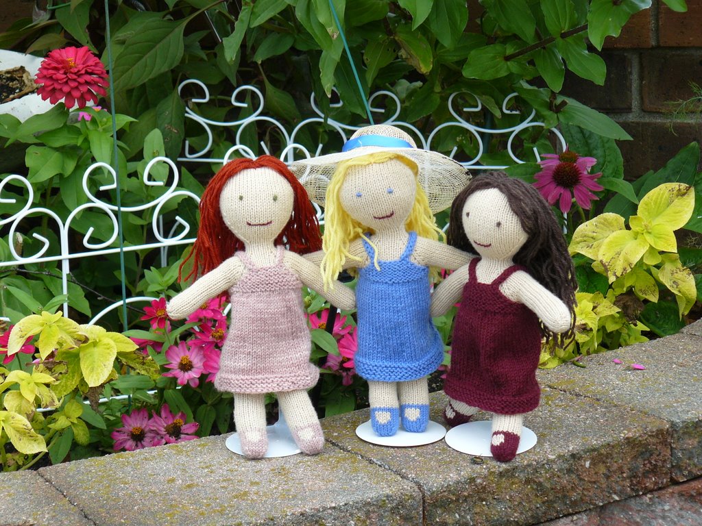 Three hand-knit dolls stand next to a small garden.